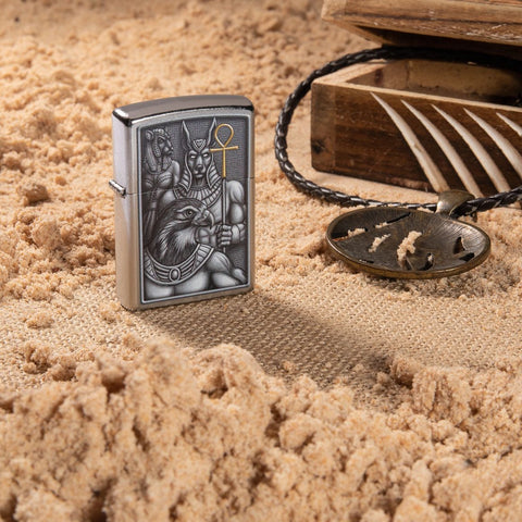 Lifestyle image of Egyptian Gods Design Street Chrome™ Windproof Lighter standing in the sand with a box and necklace next to it