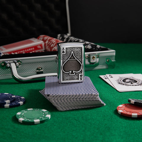 Lifestyle image of Ace Of Spades Emblem Brushed Chrome Windproof Lighter standing on a deck of cards on a poker table, with poker chips scattered around.