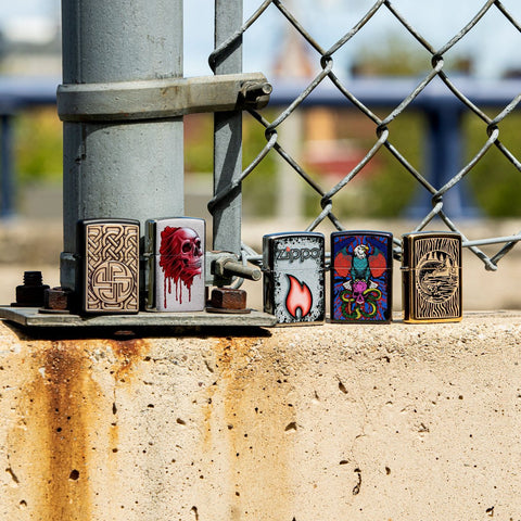 Lifestyle image of Fisherman Design High Polish Brass Windproof Lighter standing with four other lighters in front of a chain link fence