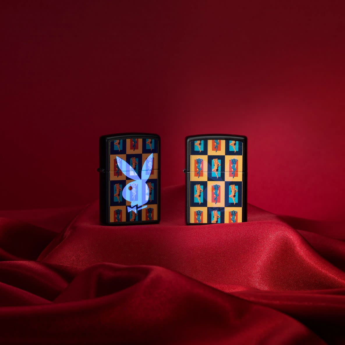 Lifestyle image of two Playboy Black Light Rabbit Head Black Matte Windproof Lighters standing on red velvet. One lighter is shown without the black light design while the other design shows the hidden black light rabbit head design.